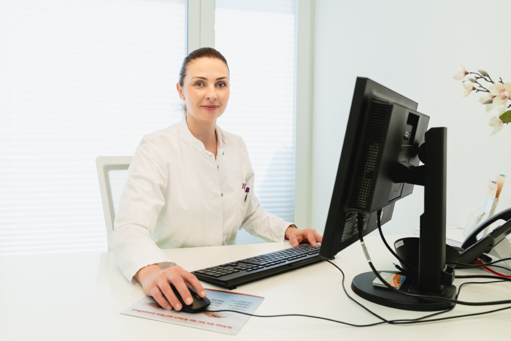 A woman in a white lab coat sits at a desk using a computer. She is looking at the camera and smiling. The office is bright and modern with a window and flowers visible in the background.