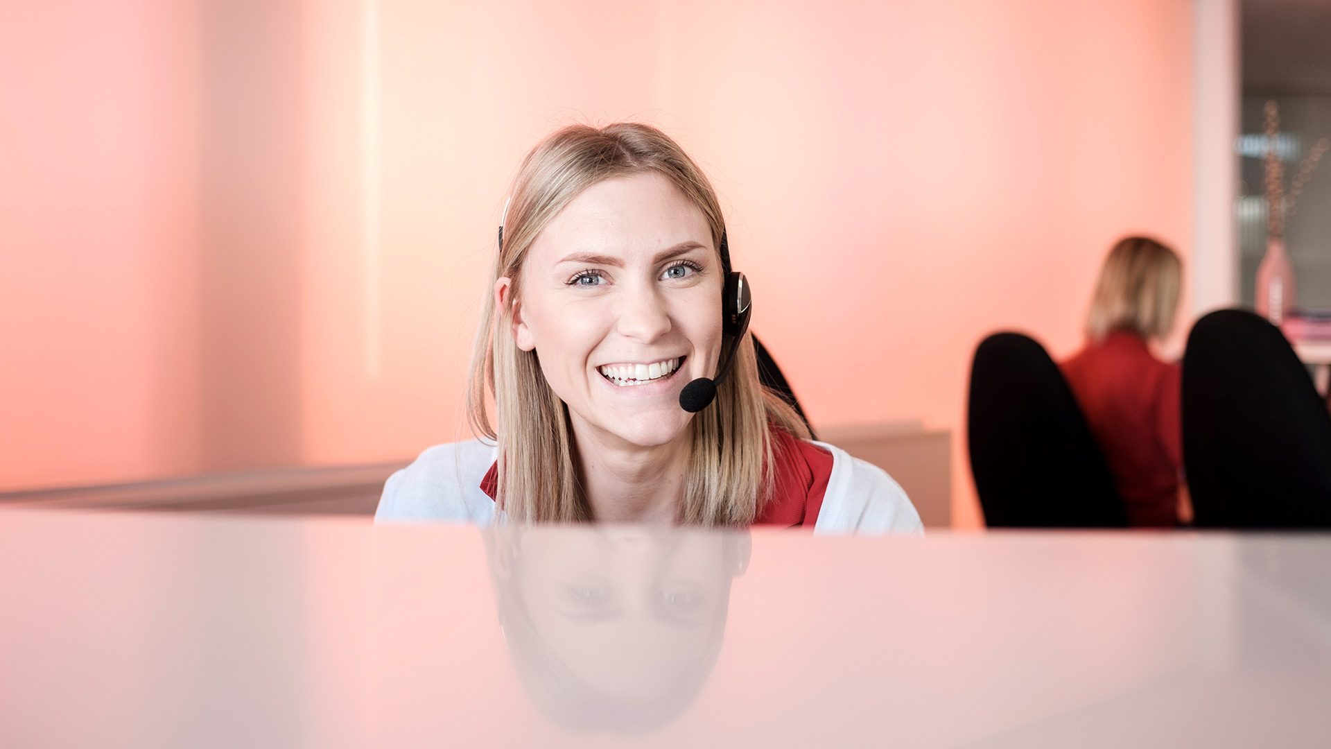 Lächelnde Mitarbeiterin mit Headset sitzt am Schreibtisch und blickt in die Kamera; im Hintergrund ist eine weitere Person zu sehen. Die freundliche Szene in einem Büro mit rosa-orangefarbenem Wand vermittelt die patientenorientierte Betreuung und Erreichbarkeit von RADIO-LOG, einem bayerischen Gesundheitsdienstleister für Radiologie, Strahlentherapie, Allgemeinmedizin, Gynäkologie und Pädiatrie an mehreren Standorten.