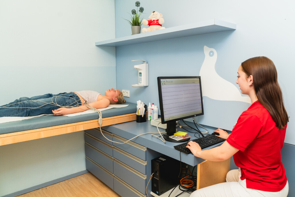 Ein Kind liegt auf einer Untersuchungsliege mit angeschlossenen Sensoren, während eine medizinische Fachkraft am Computer Messdaten überwacht. Die Szene zeigt die patientenorientierte Diagnostik und Betreuung durch ein interdisziplinäres Team bei RADIO-LOG, einem bayerischen Gesundheitsanbieter für Radiologie, Strahlentherapie, Allgemeinmedizin, Gynäkologie und Pädiatrie an mehreren Standorten.