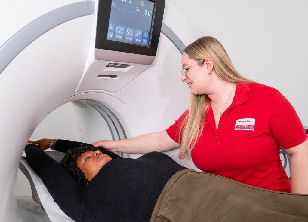 Eine Patientin liegt mit erhobenen Armen auf dem Untersuchungstisch eines CT-Scanners, während eine medizinische Fachkraft in roter Kleidung sie fürsorglich betreut und den Ablauf überwacht. Das Bild steht beispielhaft für die patientenorientierte, wohnortnahe Versorgung und interdisziplinäre Diagnostik des RADIO-LOG Teams an verschiedenen Standorten in Bayern.