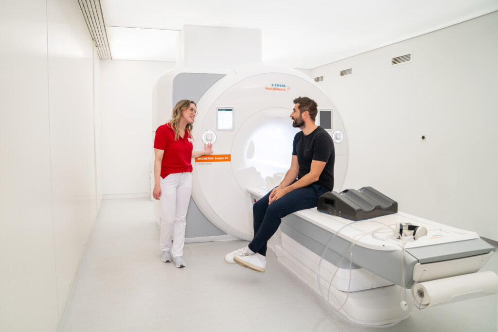 Eine Ärztin von RADIO-LOG spricht mit einem Patienten, der auf einer MRT-Untersuchungsliege in einem hell erleuchteten Behandlungsraum sitzt, und informiert ihn einfühlsam über die bevorstehende MR-Angiographie - exemplarisch für die patientenorientierte Betreuung und umfassende Diagnostik durch das interdisziplinäre Team an den RADIO-LOG Standorten in Bayern.