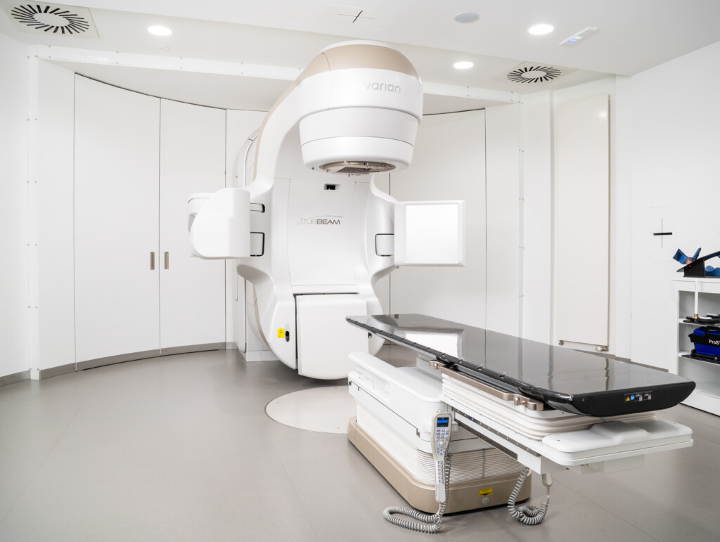 Ein moderner Linearbeschleuniger für die bildgestützte Strahlentherapie (IGRT) steht neben einer Behandlungsliege in einem hellen, gepflegten Behandlungsraum. Das Bild auf der RADIO-LOG Website veranschaulicht die fortschrittlichen technischen Möglichkeiten und das patientenorientierte Versorgungsangebot des bayerischen Gesundheitsdienstleisters im Bereich der Radiologie und Strahlentherapie.