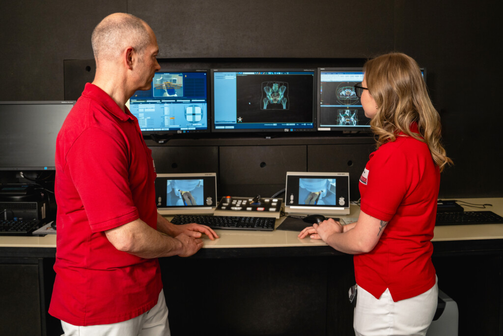 Zwei Mitarbeitende in roten Oberteilen betrachten an einer medizinischen Arbeitsstation mehrere Bildschirme mit radiologischen Aufnahmen in einer modernen Praxis für Strahlentherapie von RADIO-LOG. Das Bild unterstreicht die patientenorientierte Versorgung, interdisziplinäre Zusammenarbeit und hochwertige Diagnostik an den bayerischen Standorten des Gesundheitsanbieters.