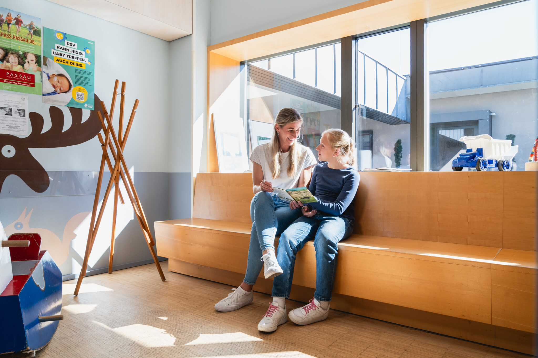 Zwei Personen sitzen gemeinsam auf einer Holzbank in einem hellen Wartebereich mit großen Fenstern und lesen in einem Buch. Im Hintergrund sind Poster sowie Kinderspielzeug zu sehen – die freundliche und einladende Atmosphäre unterstreicht das patientenorientierte Versorgungskonzept von RADIO-LOG, das an mehreren Standorten in Bayern fachübergreifende medizinische Betreuung für Patientinnen und Patienten aller Altersgruppen bietet.