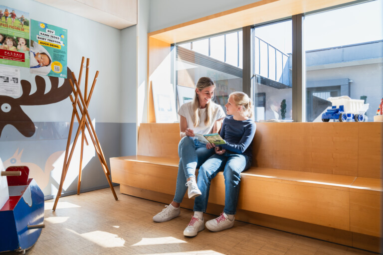 Zwei Personen sitzen gemeinsam auf einer Holzbank in einem hellen Wartebereich mit großen Fenstern und lesen in einem Buch. Im Hintergrund sind Poster sowie Kinderspielzeug zu sehen – die freundliche und einladende Atmosphäre unterstreicht das patientenorientierte Versorgungskonzept von RADIO-LOG, das an mehreren Standorten in Bayern fachübergreifende medizinische Betreuung für Patientinnen und Patienten aller Altersgruppen bietet.