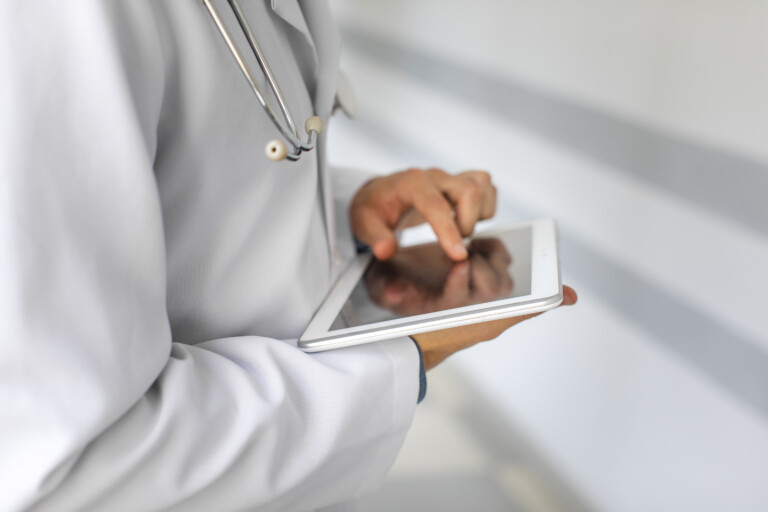A person wearing a white lab coat and stethoscope uses a tablet, likely entering or reviewing medical information. The image focuses on their hands and device, with a blurred, clinical background.