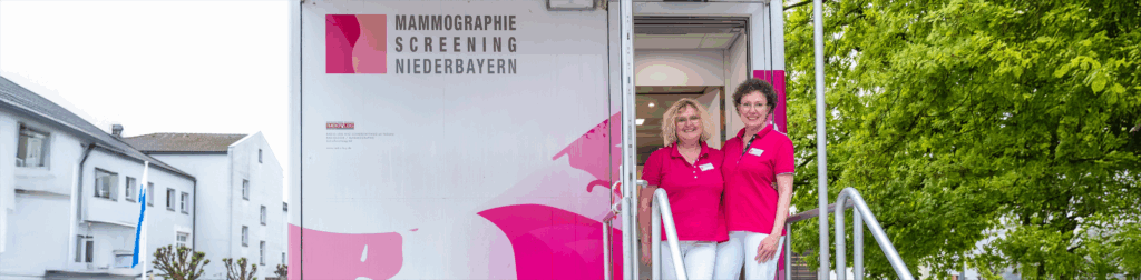 Zwei Mitarbeiterinnen in rosa Uniformen stehen am Eingang einer mobilen Mammographie-Einheit mit der Aufschrift "Mammographie-Screening Niederbayern". Die Szene verdeutlicht das wohnortnahe, patientenorientierte Versorgungsangebot und den interdisziplinären Ansatz von RADIO-LOG für moderne Diagnostik und Vorsorge in Bayern.