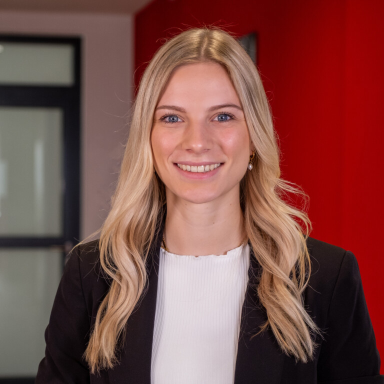Eine freundlich lächelnde Frau mit langen blonden Haaren, schwarzem Blazer und weißem Oberteil steht vor einer roten Wand und einer Glastür in einem Innenraum. Das Bild erscheint auf der RADIO-LOG Webseite, die umfassende radiologische, medizinische und therapeutische Versorgung durch interdisziplinäre Teams sowie patientenorientierte Betreuung an mehreren Standorten in Bayern bietet.