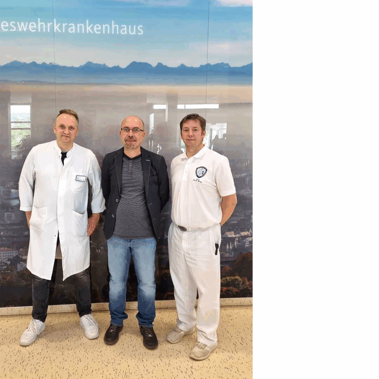 Drei Männer stehen in einem Innenraum vor einer Wand mit deutscher Aufschrift; zwei tragen weiße medizinische Arbeitskleidung, der mittlere Mann trägt Freizeitkleidung und ein dunkles Sakko. Das Foto stammt von RADIO-LOG, einem bayerischen Gesundheitsdienstleister für Radiologie, Strahlentherapie und weitere Fachbereiche, und symbolisiert einen wichtigen Fortschritt in der interdisziplinären Versorgung und wohnortnahen Behandlung von Krebspatientinnen und -patienten durch patientenzentrierte Teamarbeit.