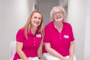 Zwei erfahrene medizinisch-technische Radiologieassistentinnen (MT-R) von RADIO-LOG begrüßen Besucher lächelnd in einer hellen Flur. Sie tragen rosa Poloshirts mit Namensschildern und repräsentieren das Mammographie-Screening der mobilen Einheit. Das Foto unterstreicht die patientenorientierte und wohnortnahe Versorgung durch das RADIO-LOG-Team in Bayern sowie das umfassende diagnostische und therapeutische Angebot aus den Bereichen Radiologie, Strahlentherapie, Allgemeinmedizin, Gynäkologie und Pädiatrie.