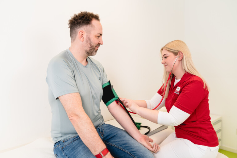 Eine medizinische Fachkraft in roter Dienstkleidung misst in einer modernen Praxisumgebung einem Patienten den Blutdruck. Das Bild veranschaulicht die patientenorientierte Betreuung, interdisziplinäre Zusammenarbeit und vielfältige Karrieremöglichkeiten bei RADIO-LOG, dem bayernweiten Gesundheitsdienstleister für Radiologie, Strahlentherapie, Allgemeinmedizin, Gynäkologie und Pädiatrie.