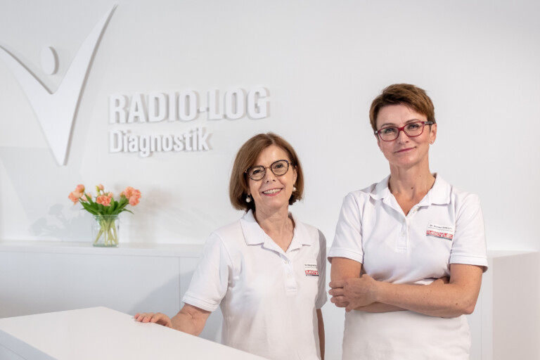 Zwei Mitarbeiterinnen in weißen Kitteln begrüßen Patientinnen an einem Empfangstresen der RADIO-LOG Praxis. Im Hintergrund ist das RADIO-LOG Diagnostik-Schild und Blumenschmuck zu sehen. Das Bild veranschaulicht die patientenorientierte Betreuung und freundliche Atmosphäre bei RADIO-LOG, einem Gesundheitsdienstleister in Bayern mit fachübergreifenden Angeboten in Radiologie, Strahlentherapie, Allgemeinmedizin, Gynäkologie und Pädiatrie an mehreren Standorten.