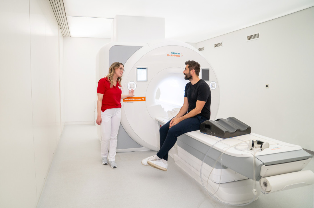 Eine Ärztin von RADIO-LOG spricht mit einem Patienten, der auf einer MRT-Untersuchungsliege in einem hell erleuchteten Behandlungsraum sitzt, und informiert ihn einfühlsam über die bevorstehende MR-Angiographie - exemplarisch für die patientenorientierte Betreuung und umfassende Diagnostik durch das interdisziplinäre Team an den RADIO-LOG Standorten in Bayern.