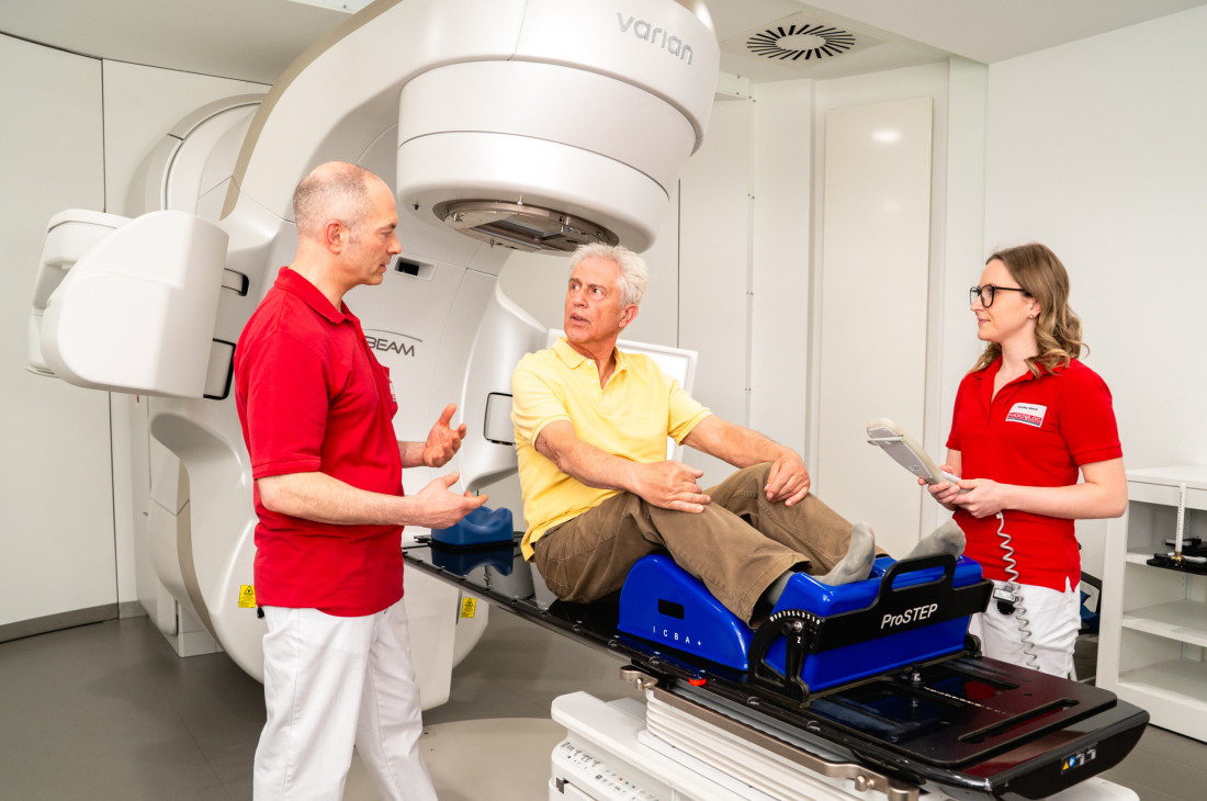Ein älterer Patient sitzt in einem Behandlungsraum von RADIO-LOG auf einer Untersuchungsliege. Zwei medizinische Fachkräfte stehen unterstützend an seiner Seite, neben einem großen Gerät für die Strahlentherapie. Das Bild vermittelt das patientenzentrierte, fachübergreifende Versorgungsangebot und die moderne Medizintechnik bei RADIO-LOG in Bayern.