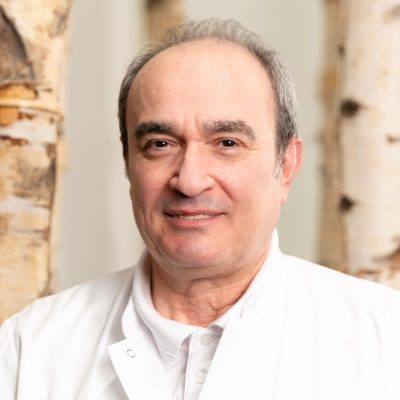 Portrait Juniku Dr. Nderim Juniku, med. univ., ein sympathisch lächelnder Facharzt im weißen Kittel, steht vor Birkenstämmen im Innenbereich - Porträt für die RADIO-LOG Website, einem bayernweiten Gesundheitsdienstleister mit Schwerpunkt auf patientenorientierter Versorgung durch interdisziplinäre Teams in Radiologie, Strahlentherapie, Allgemeinmedizin, Gynäkologie und Pädiatrie.