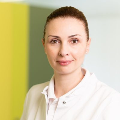 Portrait_Dr_Figurek Dr. med. Stasa Figurek von der Universität Belgrad trägt einen weißen Kittel und blickt mit neutralem Ausdruck in die Kamera. Das Foto, aufgenommen vor einem sanft verschwommenen gelben und weißen Hintergrund, stellt sie als Mitglied eines vielfältigen medizinischen Teams vor, das sich bei RADIO-LOG in Bayern für patientenzentrierte und umfassende Diagnostik sowie Behandlung im Bereich Radiologie, Strahlentherapie, Allgemeinmedizin, Gynäkologie und Pädiatrie engagiert.